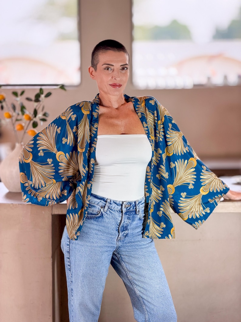May include: A blue kimono-style jacket with a gold and white floral pattern. The jacket is worn over a white tube top and light blue jeans. The sleeves are wide and the fabric appears to be lightweight.