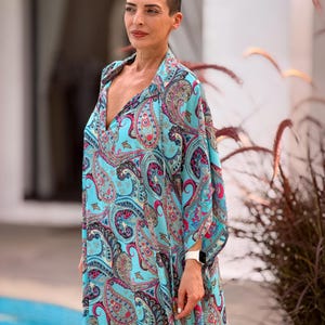 May include: A flowing, turquoise paisley print dress with a deep V-neck. The dress features a vibrant pattern of pink, blue, and gold paisley designs. The sleeves are wide and flowing. The dress is worn by a person with short hair.