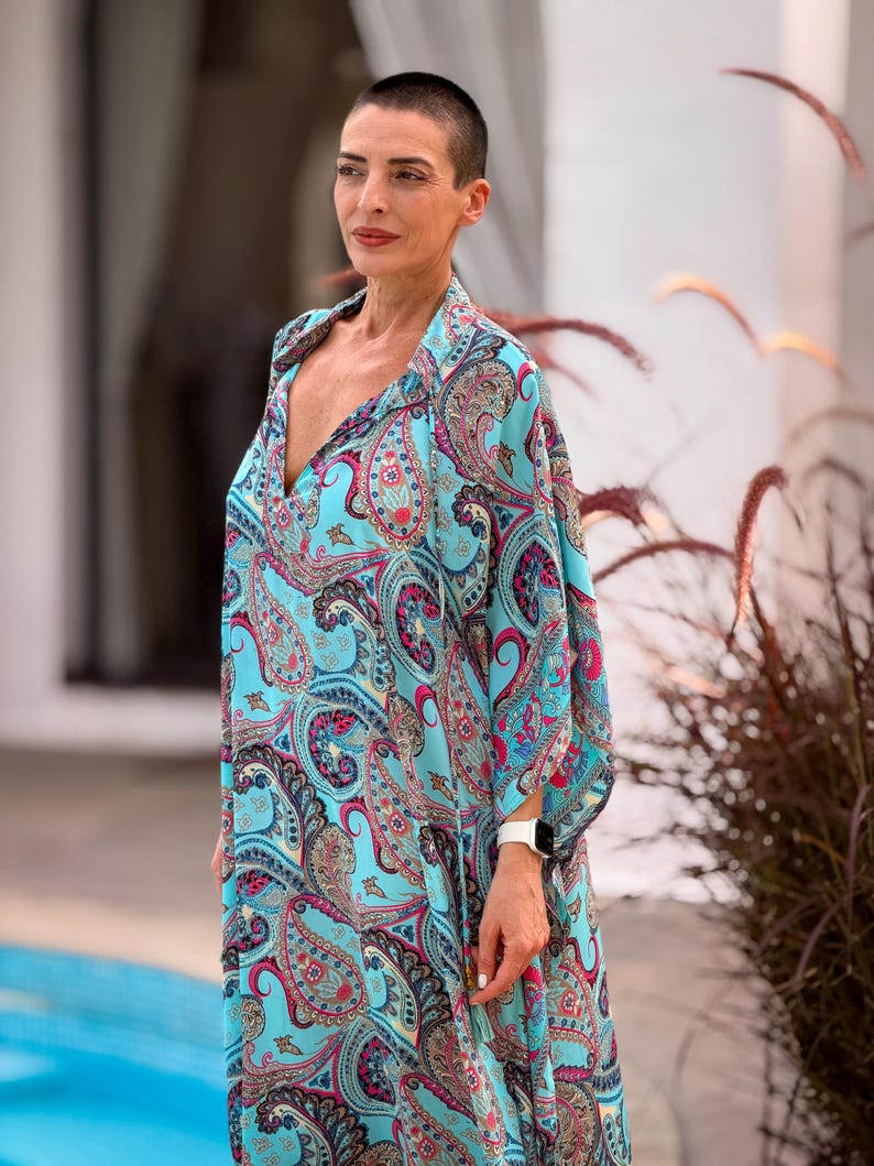 May include: A flowing, turquoise paisley print dress with a deep V-neck. The dress features a vibrant pattern of pink, blue, and gold paisley designs. The sleeves are wide and flowing. The dress is worn by a person with short hair.