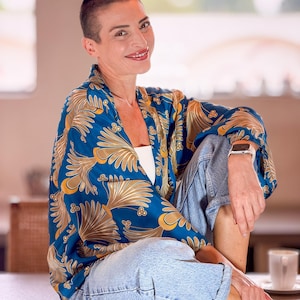 May include: A blue kimono-style top with a gold floral pattern. The top is worn with light blue denim jeans. The person is wearing a white watch and a white top. The background is blurred.