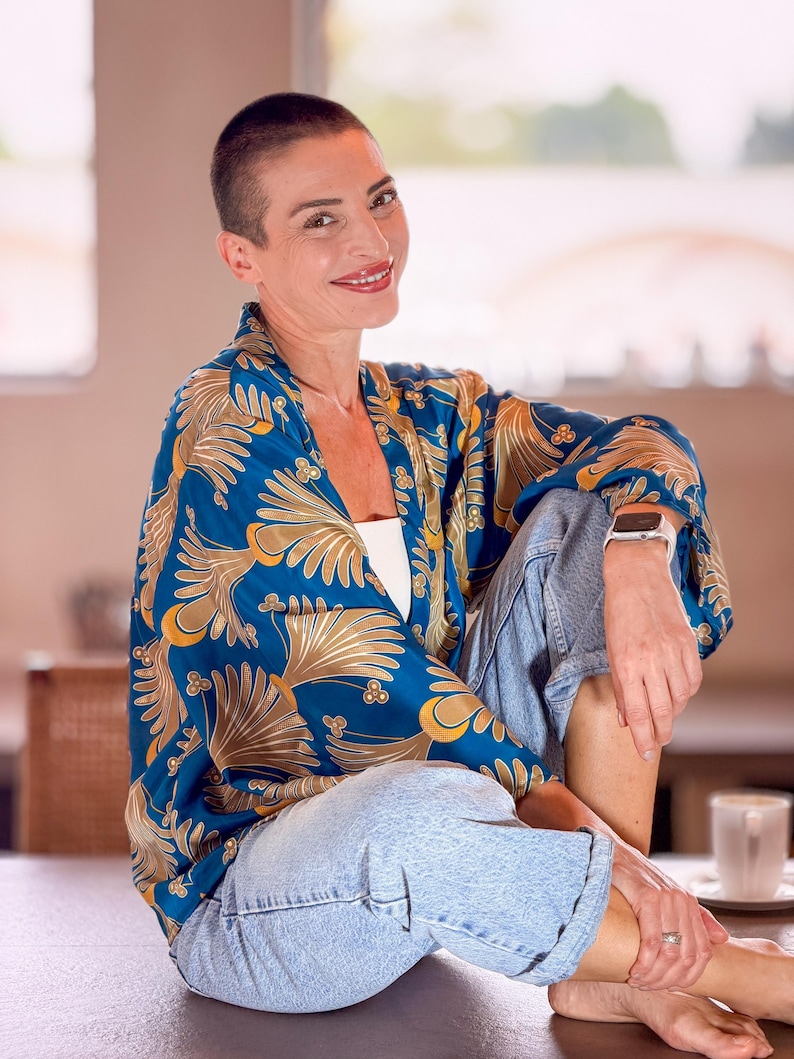 May include: A blue kimono-style top with a gold floral pattern. The top is worn with light blue denim jeans. The person is wearing a white watch and a white top. The background is blurred.