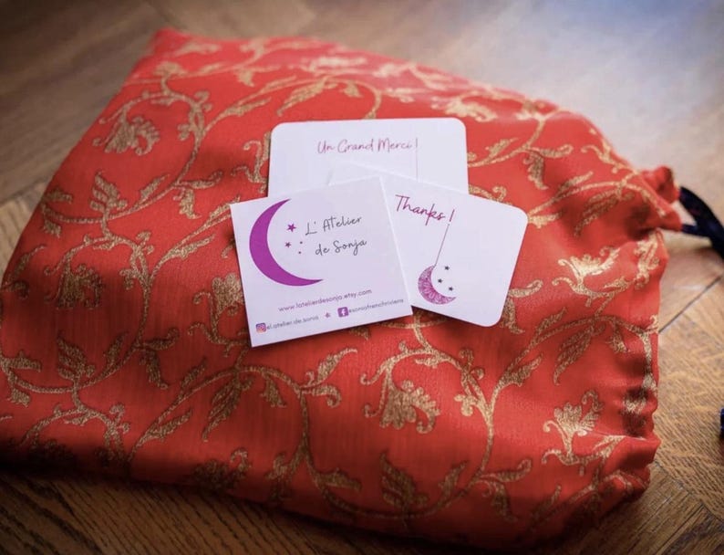 May include: A red drawstring bag with a gold floral pattern, accompanied by three small white cards. The cards feature text and a purple crescent moon design. One card says "Thanks!" and another says "Un Grand Merci!"