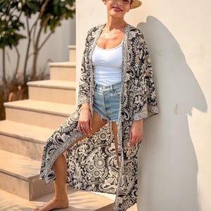 May include: A woman wearing a straw fedora hat with a black band, a white tank top, and denim shorts. She is also wearing a long, open black and white patterned kimono with a silver trim. The woman is standing on a set of outdoor stairs.