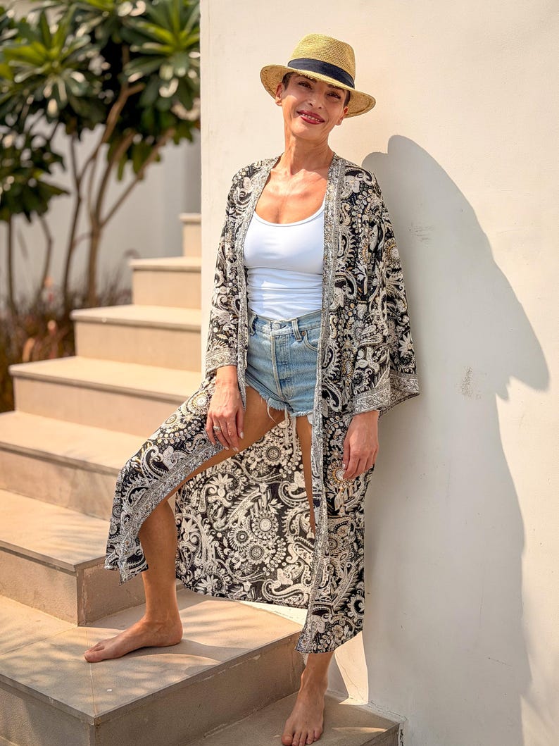 May include: A woman wearing a straw fedora hat with a black band, a white tank top, and denim shorts. She is also wearing a long, open black and white patterned kimono with a silver trim. The woman is standing on a set of outdoor stairs.