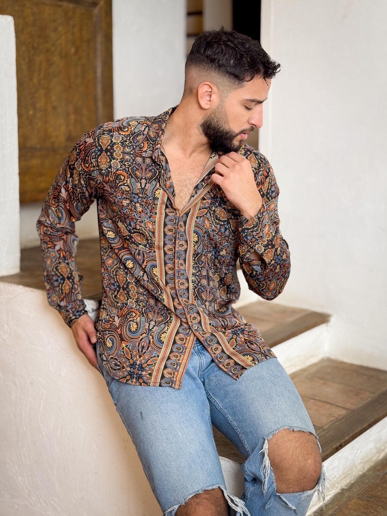 May include: A man wearing a long-sleeved shirt with a paisley pattern in shades of brown, blue, and orange. He is wearing blue jeans with a distressed knee.