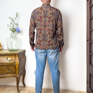 May include: A man wearing a long-sleeved shirt with a colorful paisley pattern and blue jeans.