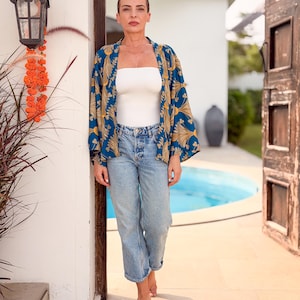 May include: A woman wearing a blue and gold patterned kimono-style jacket, a white tube top, and light blue jeans. The woman is standing in a doorway with a pool visible in the background. An outdoor lamp and orange flowers are on the wall.