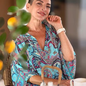 May include: A woman wearing a turquoise paisley print top with a deep V-neck. She is smiling and wearing a white watch. A white teapot and teacup are on the table in front of her. The background is blurred.