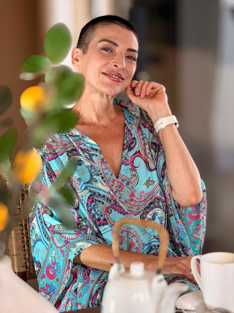May include: A woman wearing a turquoise paisley print top with a deep V-neck. She is smiling and wearing a white watch. A white teapot and teacup are on the table in front of her. The background is blurred.