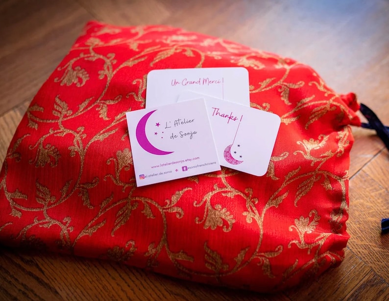 May include: A red fabric pouch with gold floral embroidery and a white card with the text "Un Grand Merci!" and a white card with the text "Thanks!" and a business card with the text "L'Atelier de Sonja" and a website address.
