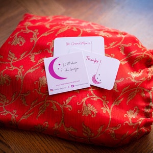 May include: A red fabric bag with gold floral embroidery and a blue drawstring. The bag is sitting on a wooden surface with three white cards. The cards have the text "Un Grand Merci!" and "L'Atelier de Sonya" with a pink crescent moon and stars. The third card says "Thanks!" with a crescent moon and stars.