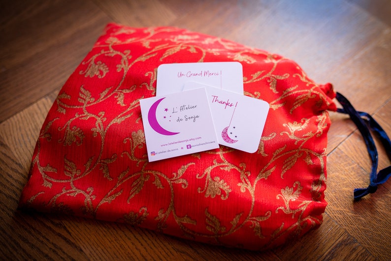 May include: A red fabric bag with gold floral embroidery and a blue drawstring. The bag is sitting on a wooden surface with three white cards. The cards have the text "Un Grand Merci!" and "L'Atelier de Sonya" with a pink crescent moon and stars. The third card says "Thanks!" with a crescent moon and stars.