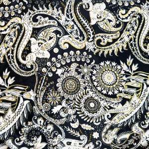 May include: Close-up of a fabric with a black background and a repeating paisley pattern. The pattern features white and gold floral and swirl designs, creating a detailed and ornate textile.