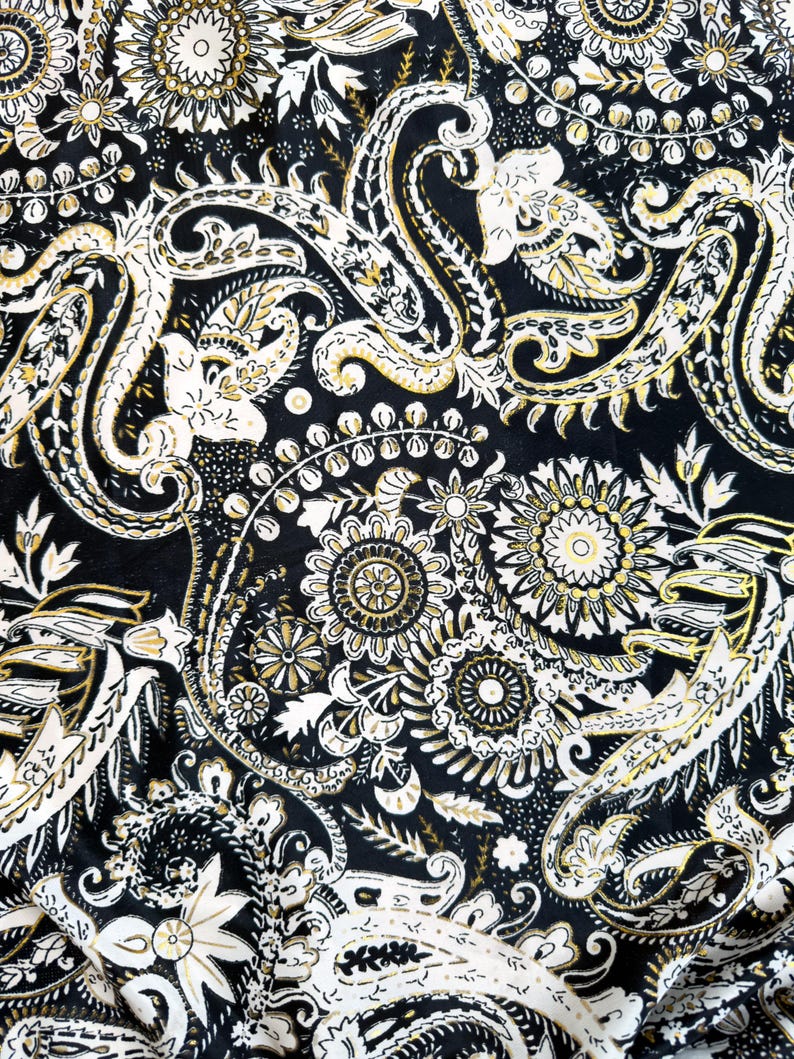 May include: Close-up of a fabric with a black background and a repeating paisley pattern. The pattern features white and gold floral and swirl designs, creating a detailed and ornate textile.