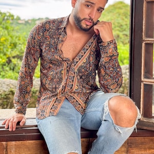 May include: A man wearing a long-sleeved paisley patterned shirt with a brown, orange, and blue color scheme. He is wearing light blue jeans with rips in the knees and black shoes.