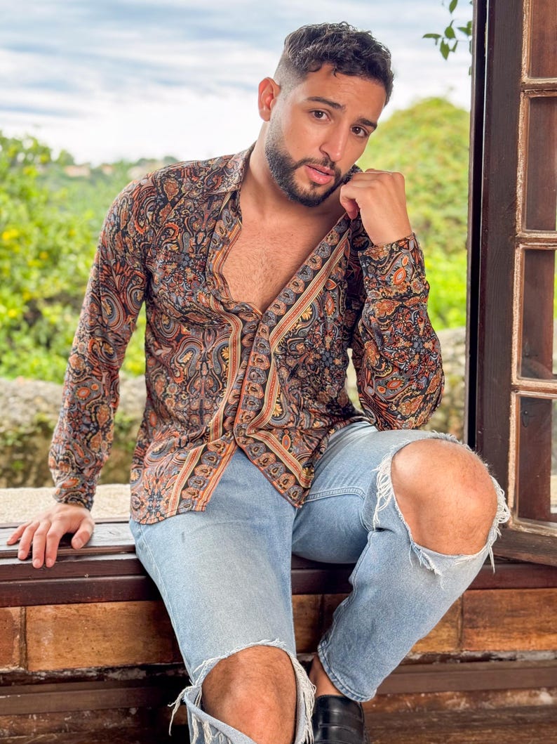 May include: A man wearing a long-sleeved paisley patterned shirt with a brown, orange, and blue color scheme. He is wearing light blue jeans with rips in the knees and black shoes.