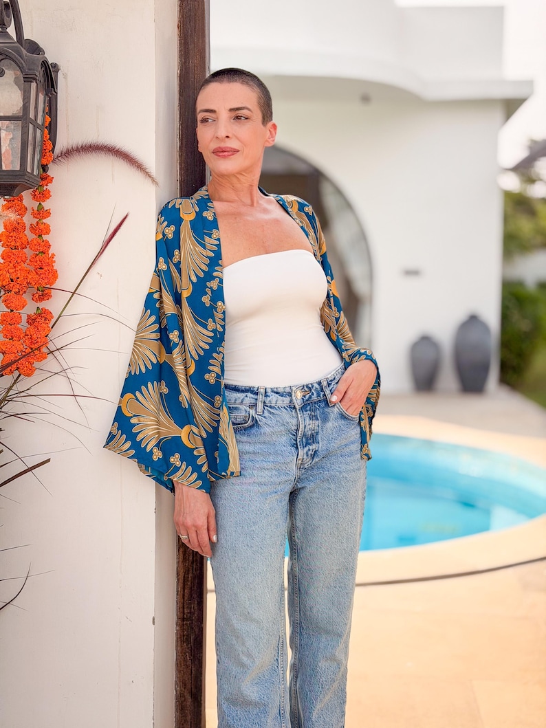 May include: A woman wearing a blue and gold patterned kimono-style jacket over a white tube top and light wash denim jeans. The jacket features a botanical design. The background includes a white building, a pool, and orange flowers.