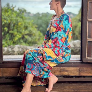 May include: A woman wearing a colourful patterned kimono-style robe sits on a wooden bench, looking out of a window. The robe has a mix of red, blue, yellow, and green colours.
