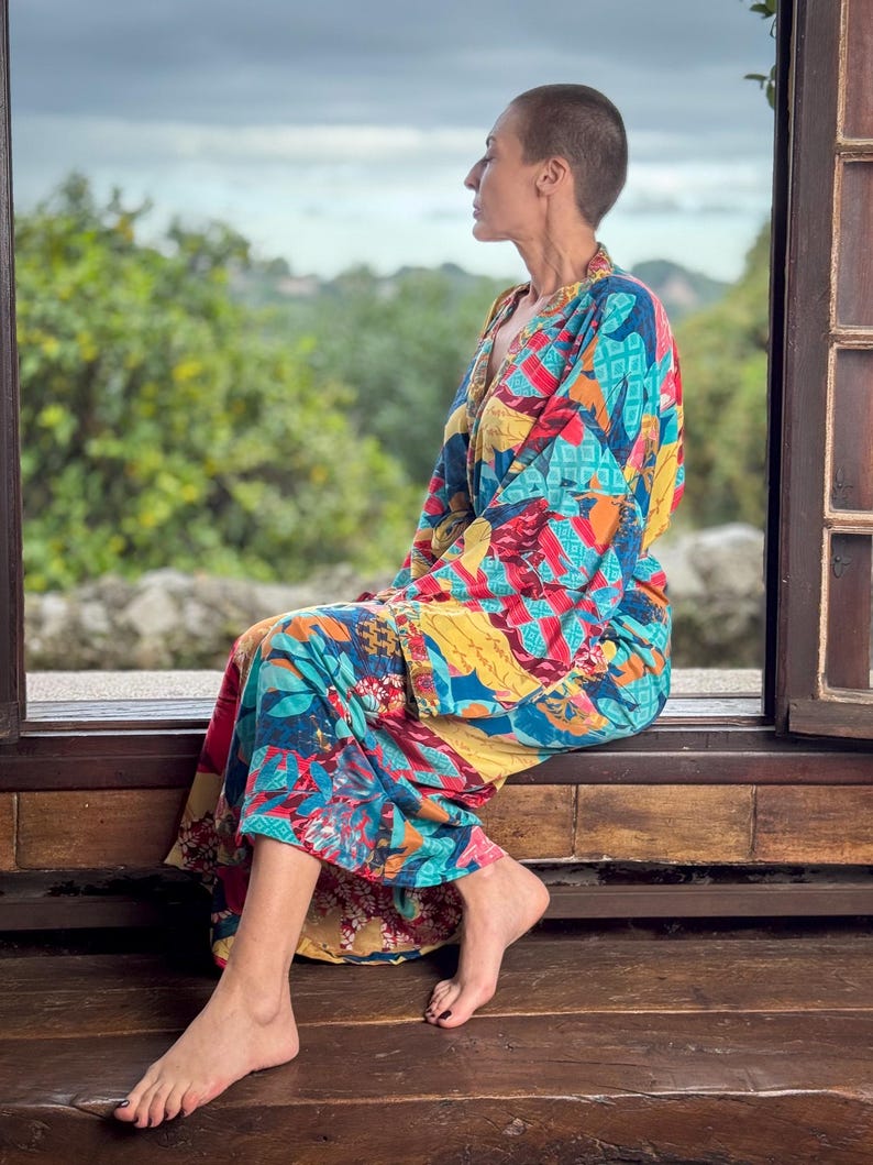May include: A woman wearing a colourful patterned kimono-style robe sits on a wooden bench, looking out of a window. The robe has a mix of red, blue, yellow, and green colours.