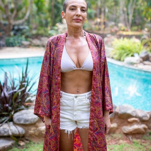 Peut inclure: Une femme portant un kimono &agrave; motifs rouge et or avec un haut de bikini blanc et un short en jean blanc. Elle se tient devant une piscine.