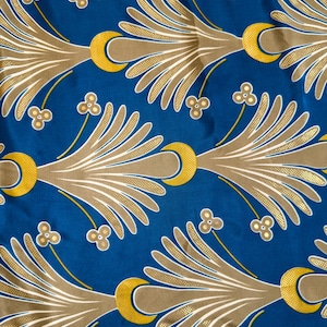May include: A close-up of a blue fabric with a repeating pattern. The pattern features gold and tan stylized leaf-like shapes, yellow crescent moons, and small floral accents. The design is symmetrical and creates a decorative textile.