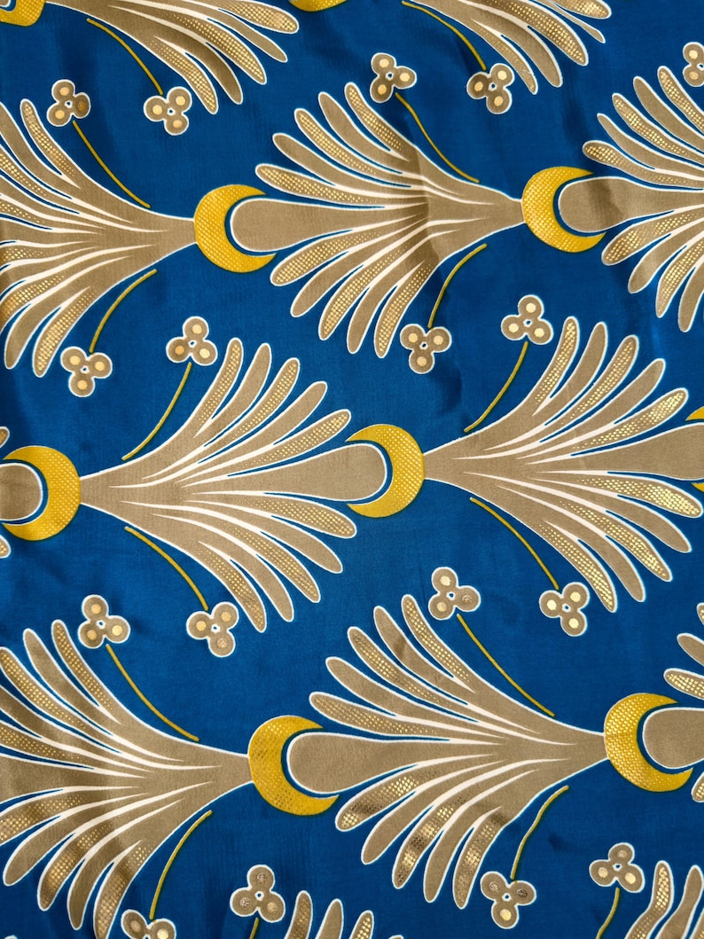 May include: A close-up of a blue fabric with a repeating pattern. The pattern features gold and tan stylized leaf-like shapes, yellow crescent moons, and small floral accents. The design is symmetrical and creates a decorative textile.