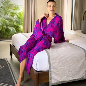 May include: A vibrant, flowing robe in shades of fuchsia and purple, featuring a bold floral pattern. The robe is worn open, revealing a glimpse of the wearer. The scene is set in a bedroom with a bed and a window.