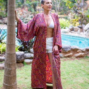 Peut inclure: Une femme portant un long kimono rouge &agrave; motifs paisley avec un haut de bikini blanc en dessous. Elle se tient dans un jardin avec un palmier derri&egrave;re elle.