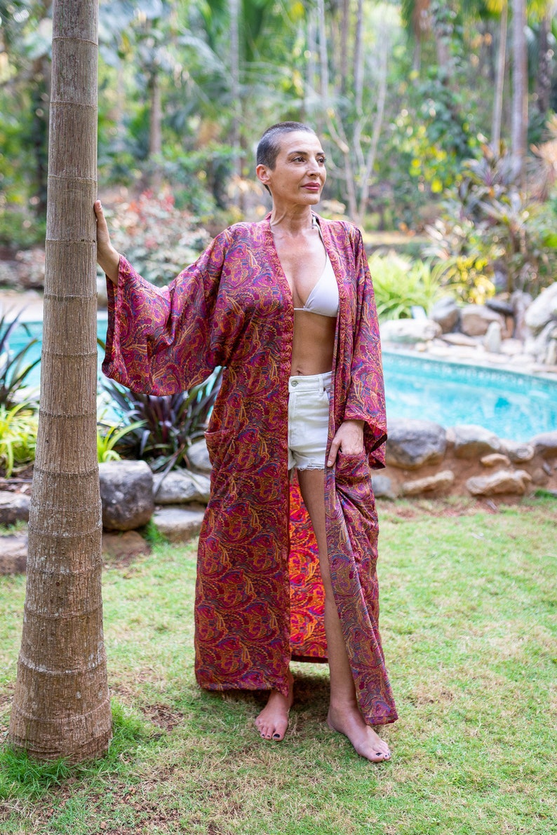 Peut inclure: Une femme portant un long kimono rouge &agrave; motifs paisley avec un haut de bikini blanc en dessous. Elle se tient dans un jardin avec un palmier derri&egrave;re elle.