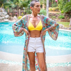 May include: A woman wearing a white bikini top and white denim shorts with a patterned kimono over top. She is standing by a swimming pool.