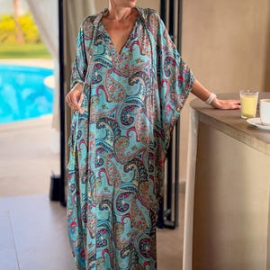 May include: A flowing, turquoise paisley print caftan dress with a V-neck and wide sleeves. The dress features a colorful pattern of red, pink, and blue paisley designs. A tassel detail hangs from the neckline. The dress is floor-length.