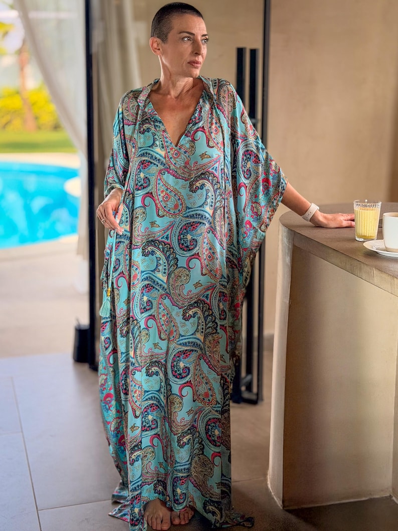 May include: A flowing, turquoise paisley print caftan dress with a V-neck and wide sleeves. The dress features a colorful pattern of red, pink, and blue paisley designs. A tassel detail hangs from the neckline. The dress is floor-length.