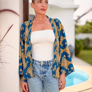May include: A woman wearing a blue kimono with a gold floral pattern over a white tube top and light blue jeans. The kimono has long sleeves and is open in the front. The woman is standing outside.