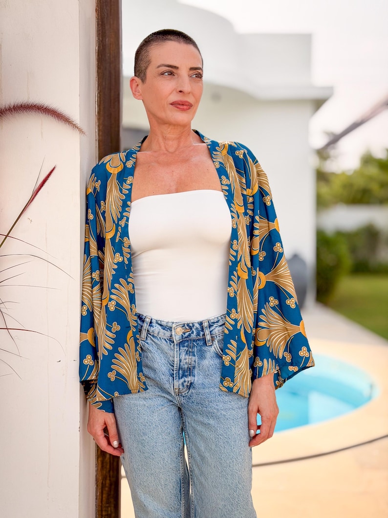 May include: A woman wearing a blue kimono with a gold floral pattern over a white tube top and light blue jeans. The kimono has long sleeves and is open in the front. The woman is standing outside.