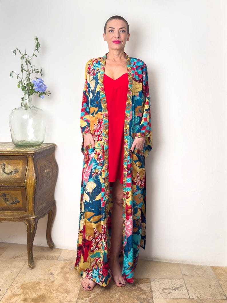 May include: A long, flowing floral print robe with a red slip dress underneath. The robe has a deep V-neckline and long sleeves. The floral print is in shades of blue, yellow, red, and green.