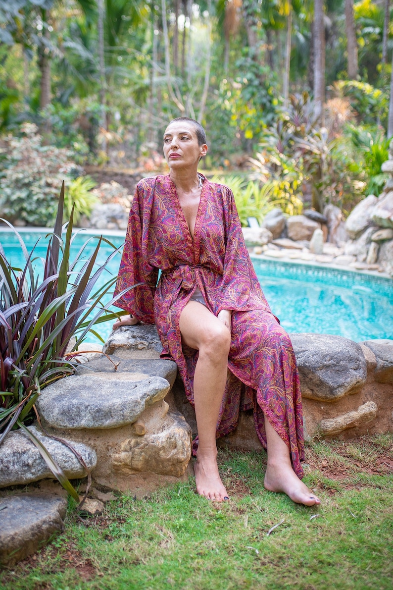 Peut inclure: Une femme portant une robe de chambre &agrave; motifs paisley rose et violet est assise sur un rocher au bord d'une piscine. La robe est ouverte et les jambes de la femme sont visibles. La piscine est en arri&egrave;re-plan et il y a des plantes vertes autour de la piscine.