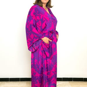 May include: A long, flowing robe in a vibrant floral print of magenta and purple. The robe features wide sleeves, a V-neck, and a matching belt. The model is wearing black sandals.