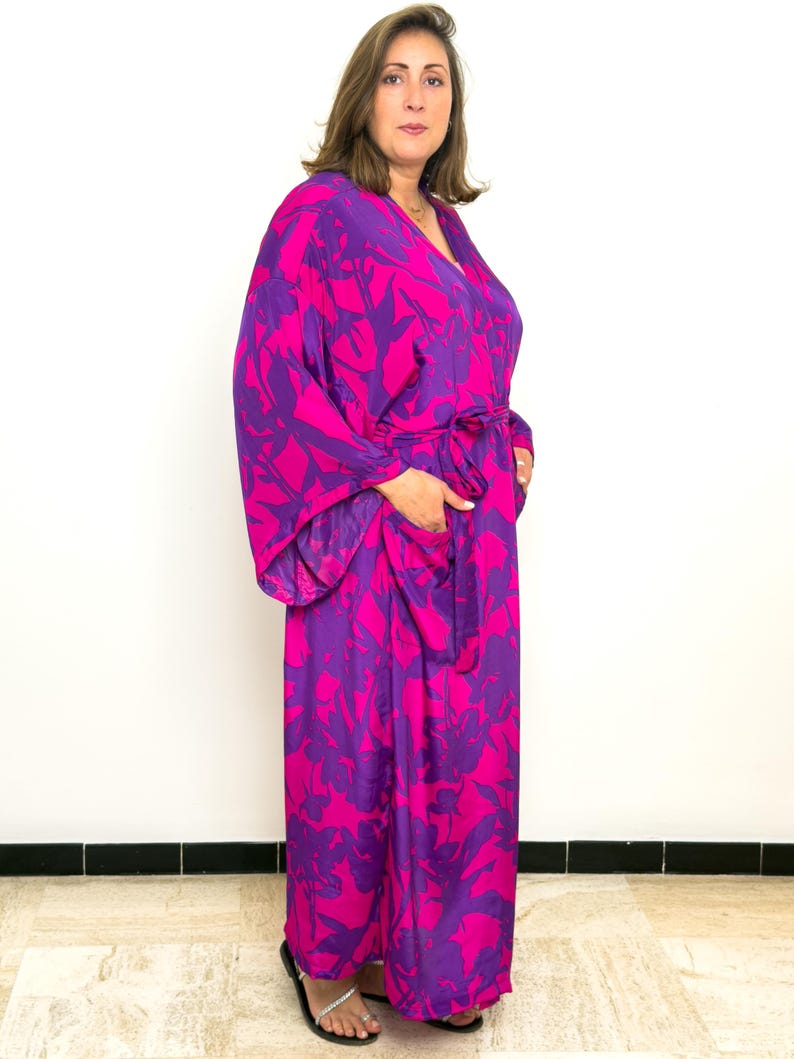 May include: A long, flowing robe in a vibrant floral print of magenta and purple. The robe features wide sleeves, a V-neck, and a matching belt. The model is wearing black sandals.