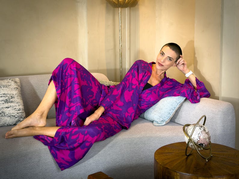 May include: A woman wearing a vibrant purple and fuchsia floral print outfit, including a kimono-style top and matching pants, reclines on a gray sofa. The outfit's bold pattern is a striking contrast against the neutral background. A decorative globe sits on a wooden table.