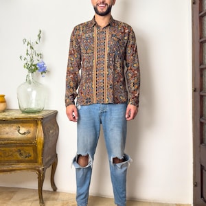 May include: A man wearing a long-sleeved shirt with a paisley pattern in shades of brown, blue, and gold. The shirt has a button-down collar and a relaxed fit. He is also wearing blue jeans with distressed knees.
