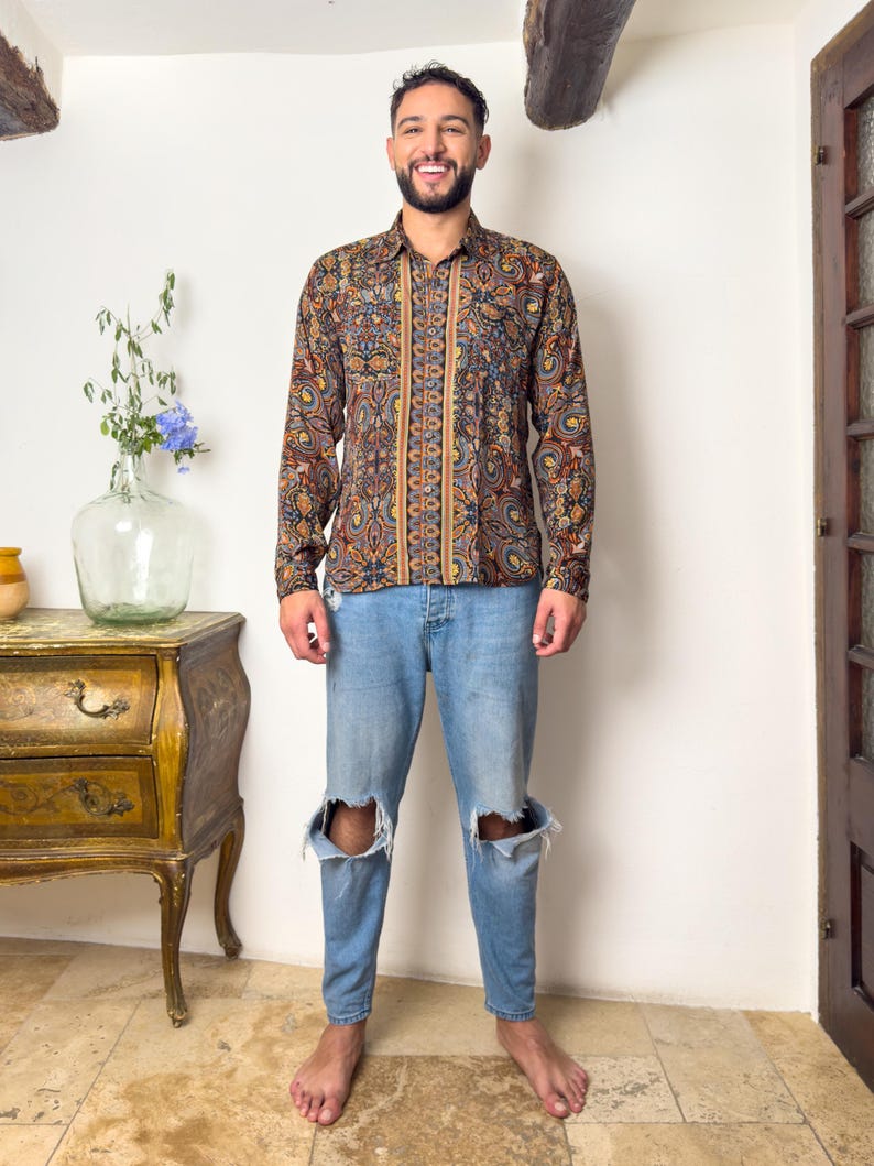 May include: A man wearing a long-sleeved shirt with a paisley pattern in shades of brown, blue, and gold. The shirt has a button-down collar and a relaxed fit. He is also wearing blue jeans with distressed knees.