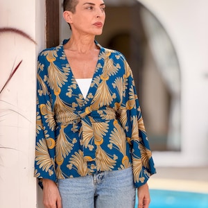 May include: A teal kimono-style top with a gold and white floral pattern. The top has long sleeves and a tie closure at the waist. The model is wearing light blue jeans and a white top underneath.
