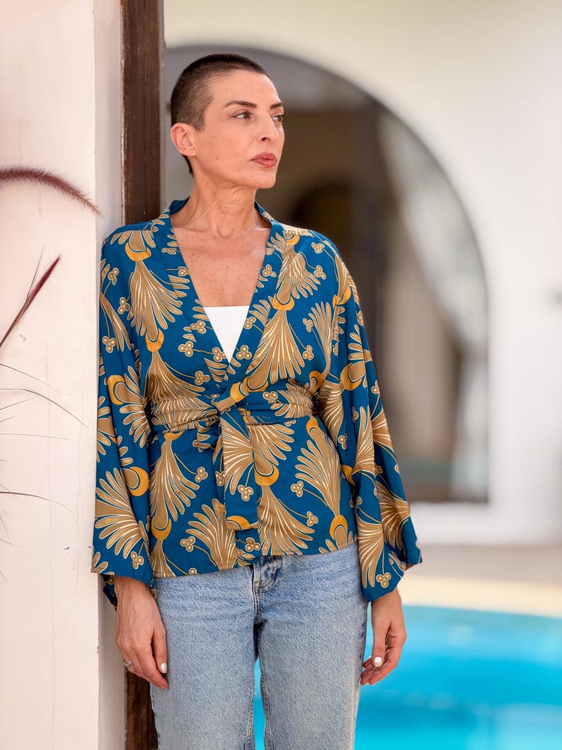 May include: A teal kimono-style top with a gold and white floral pattern. The top has long sleeves and a tie closure at the waist. The model is wearing light blue jeans and a white top underneath.