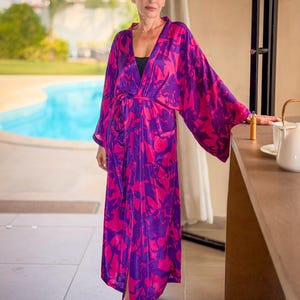 May include: A long, flowing robe in a vibrant pink and purple floral pattern. The robe has wide sleeves and a matching belt. The model is standing near a pool, wearing a black top underneath.
