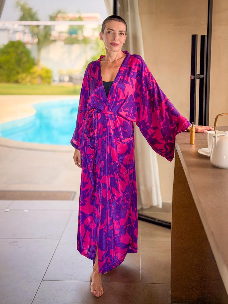 May include: A long, flowing robe in a vibrant pink and purple floral pattern. The robe has wide sleeves and a matching belt. The model is standing near a pool, wearing a black top underneath.