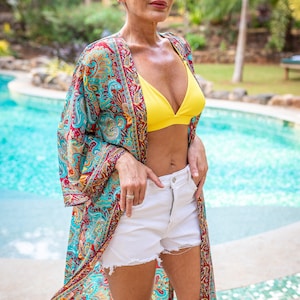 May include: A woman wearing a colorful patterned kimono over a yellow bikini top and white denim shorts. She is standing in front of a swimming pool.