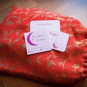 May include: A red fabric pouch with gold floral embroidery and a blue drawstring. The pouch contains two small white cards with pink and black text. The text on the cards reads "Un Grand Merci!" and "L'Atelier de Sonya" with a star and crescent moon design.