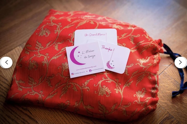 May include: A red fabric pouch with gold floral embroidery and a blue drawstring. The pouch contains two small white cards with pink and black text. The text on the cards reads "Un Grand Merci!" and "L'Atelier de Sonya" with a star and crescent moon design.