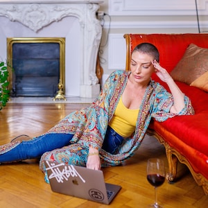 May include: A woman wearing a colorful patterned kimono sits on the floor with a laptop and a glass of red wine. The laptop has a sticker on it that says "Thissam".