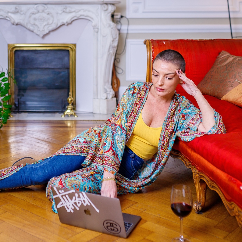 May include: A woman wearing a colorful patterned kimono sits on the floor with a laptop and a glass of red wine. The laptop has a sticker on it that says "Thissam".
